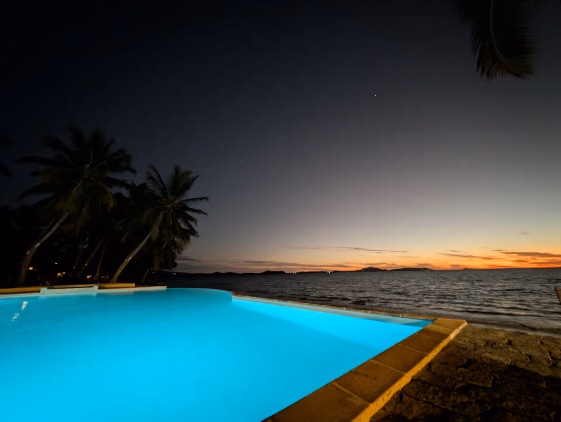 This is an image showing off the sunset over the Mozambique Channel in Nosy Be Madagascar with a landscape made of blue swimming pool and coconut palm trees.